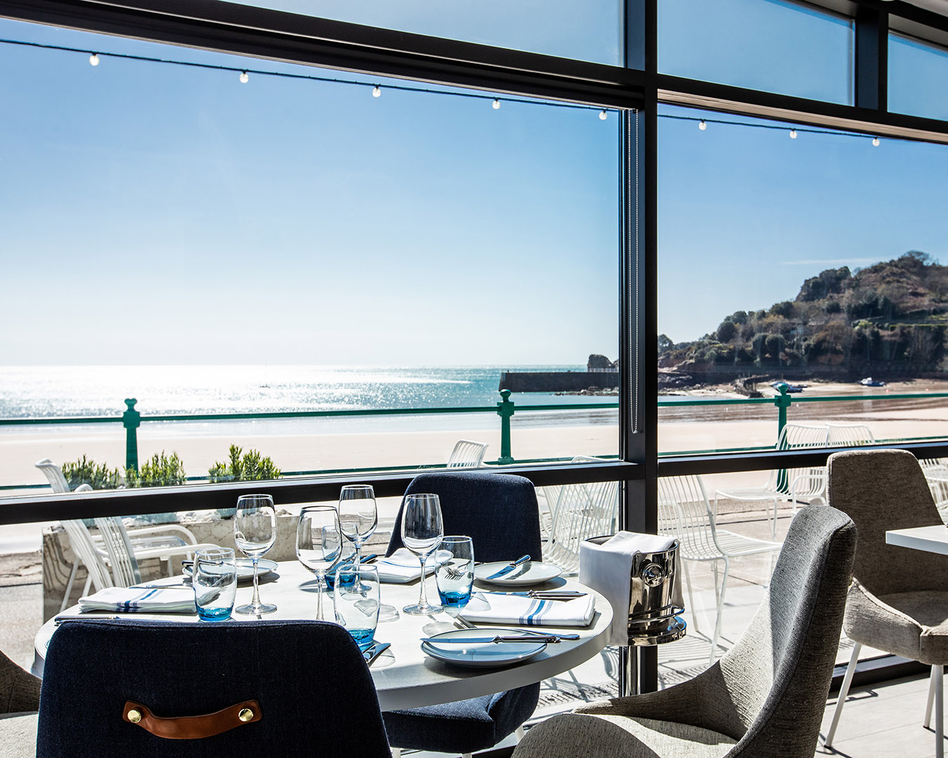 A chic restaurant interior featuring navy and sand-coloured textured upholstery and a view over a sunny beach