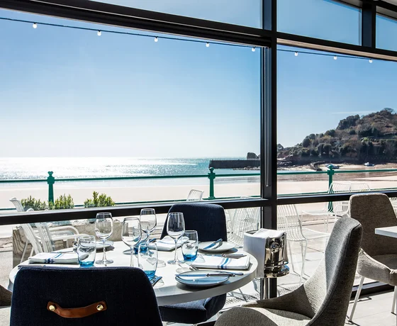 A chic restaurant interior featuring navy and sand-coloured textured upholstery and a view over a sunny beach