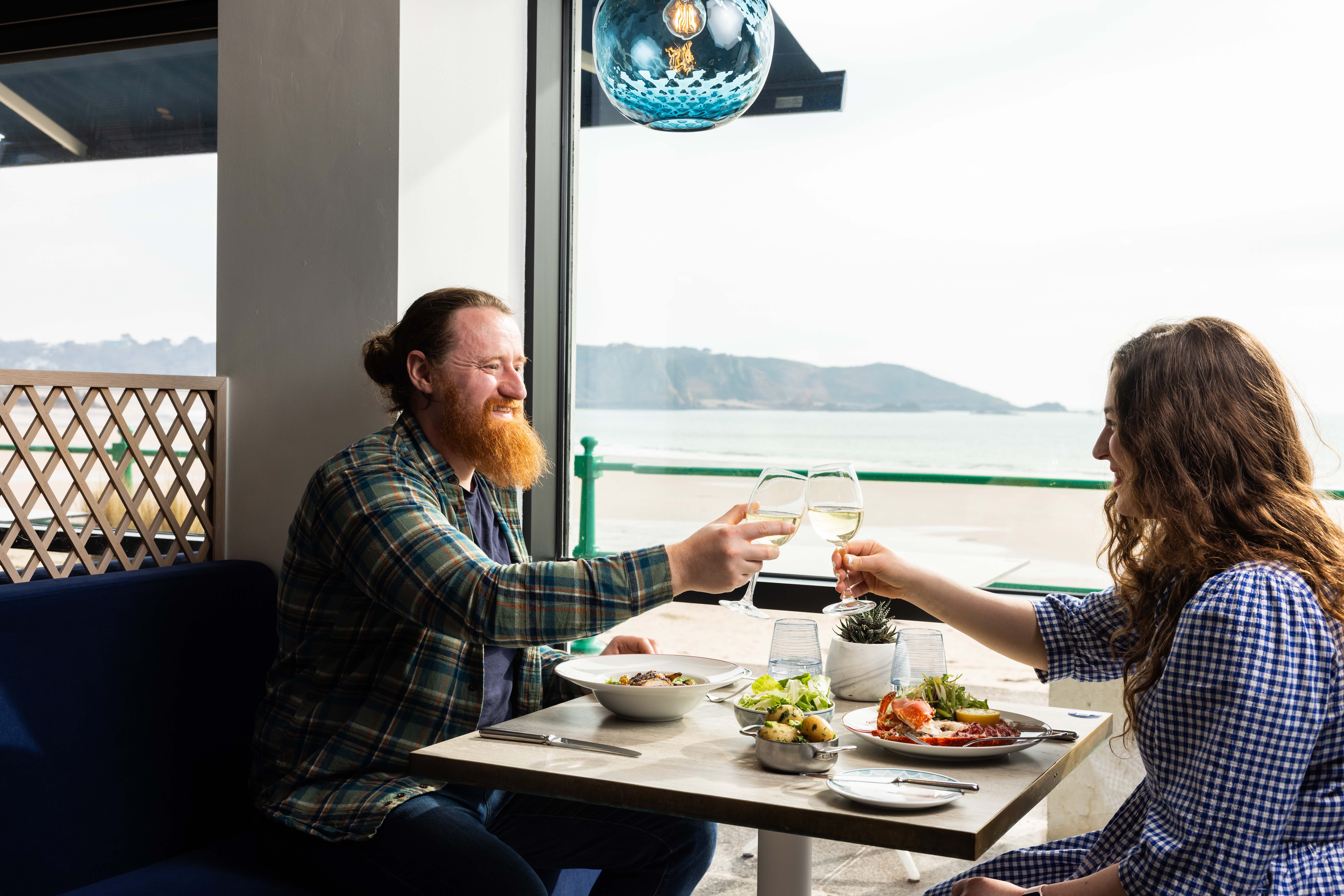A couple toast over a table of food with the view of St Brelades Bay in the background