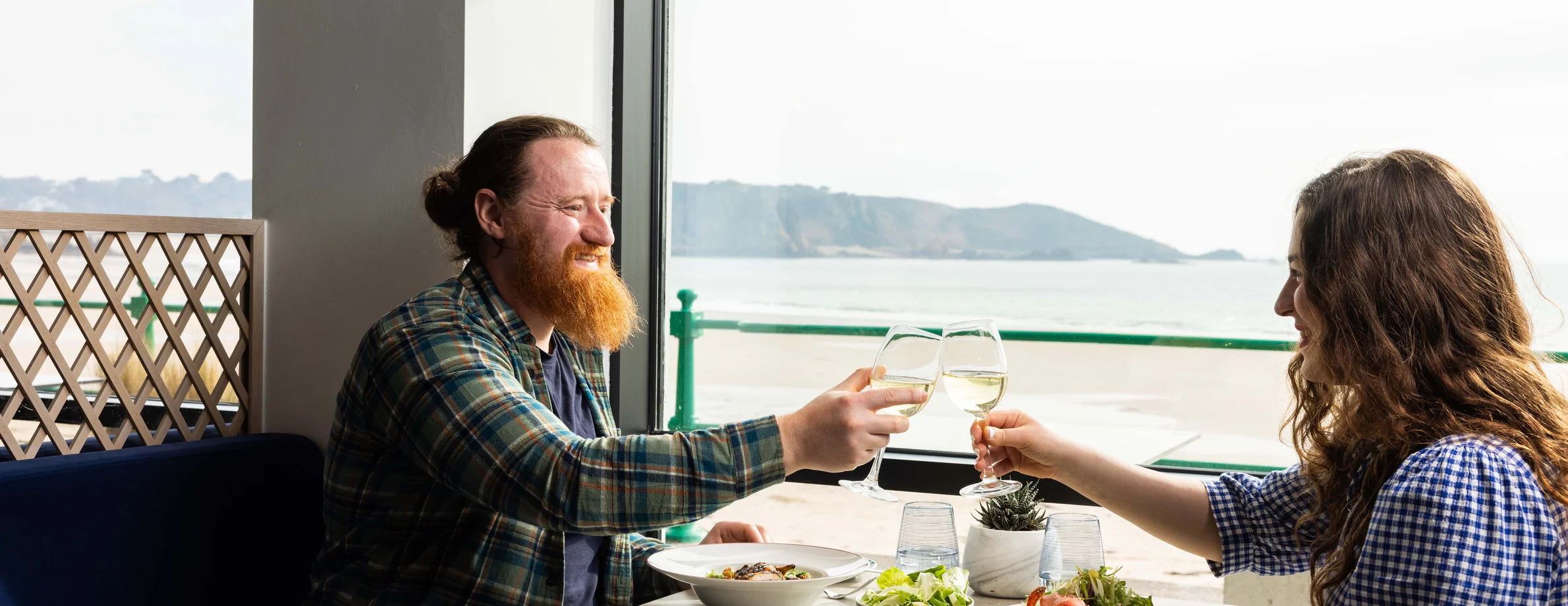 A couple toast over a table of food with the view of St Brelades Bay in the background