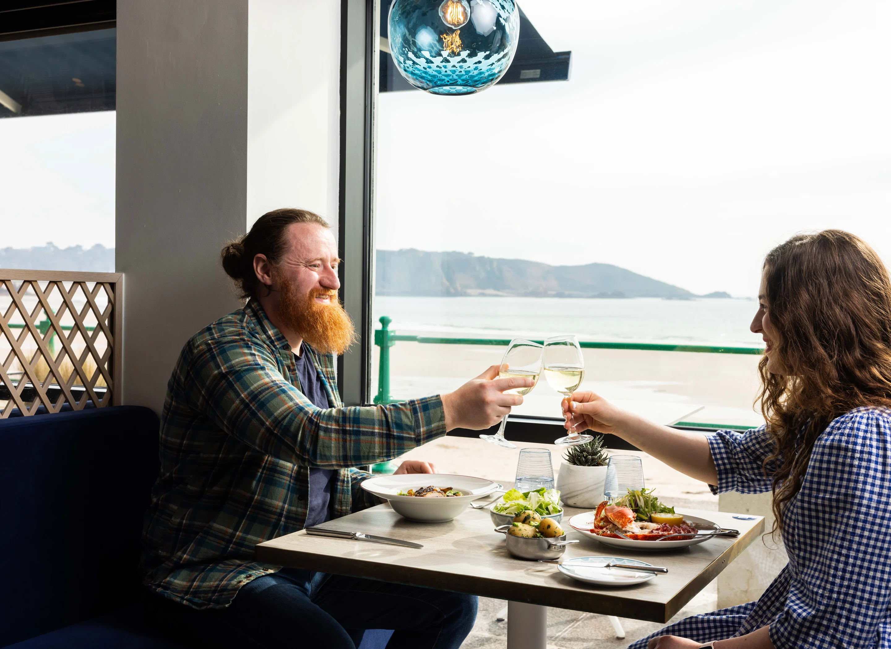 A couple toast over a table of food with the view of St Brelades Bay in the background