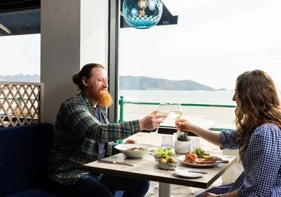 A couple toast over a table of food with the view of St Brelades Bay in the background