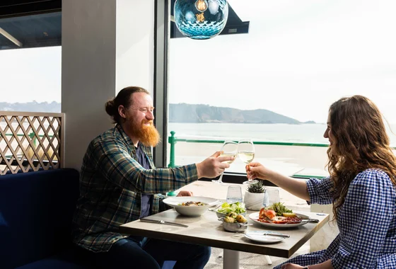 A couple toast over a table of food with the view of St Brelades Bay in the background