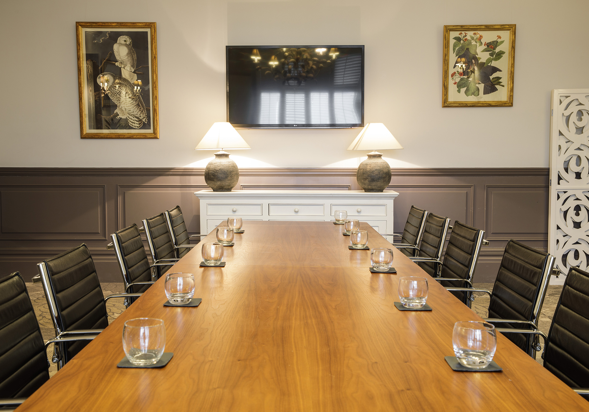 A meeting room equipped with a long boardroom table, boardroom seating and a digital screen