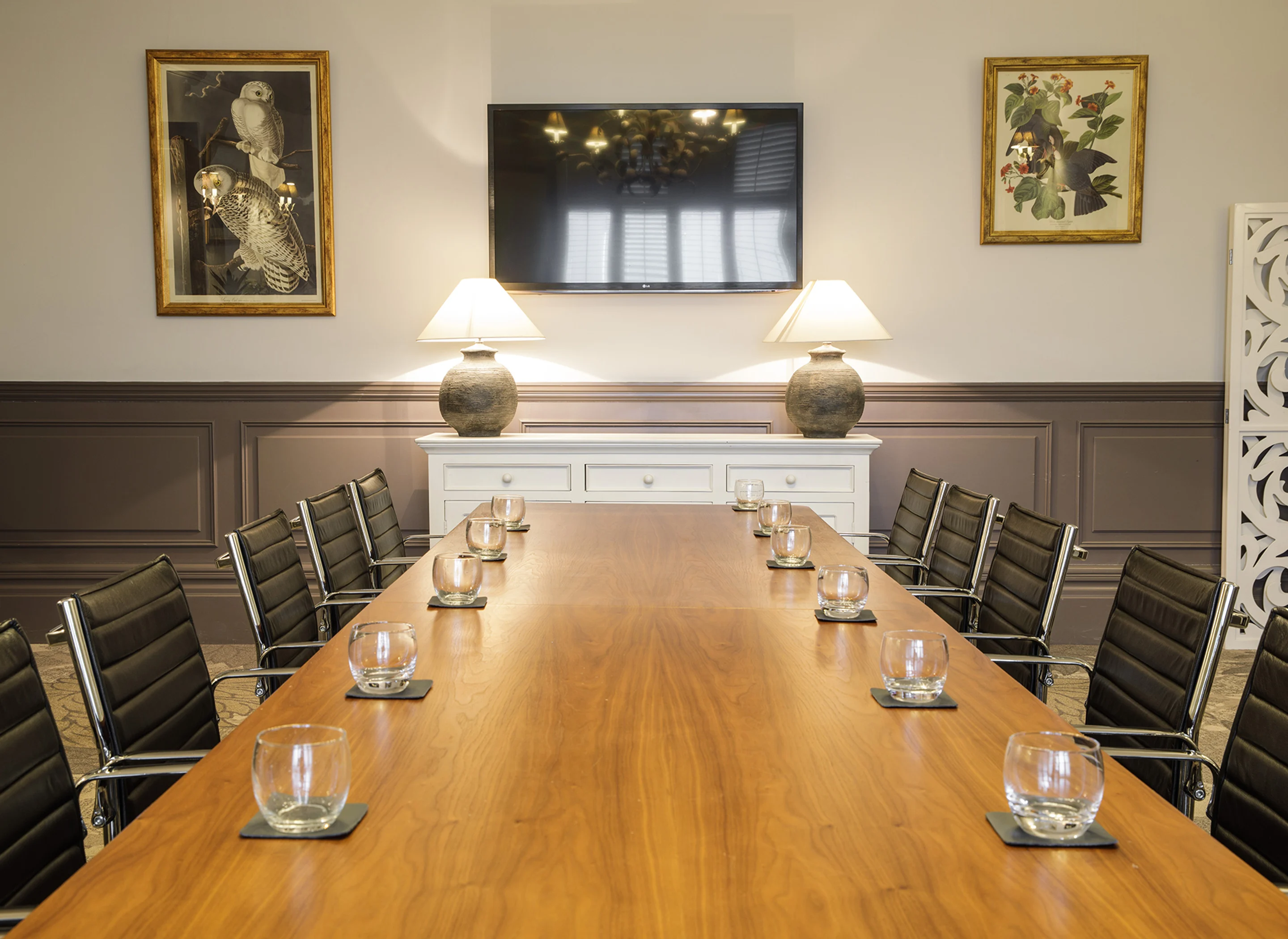 A meeting room equipped with a long boardroom table, boardroom seating and a digital screen