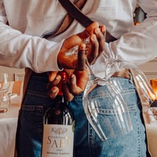 A waiter stands beside a table, holding a bottle of wine and wine glasses
