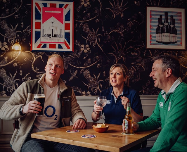 A family with an adult son watch the big game together, each holding a drink and wearing a rugby shirt